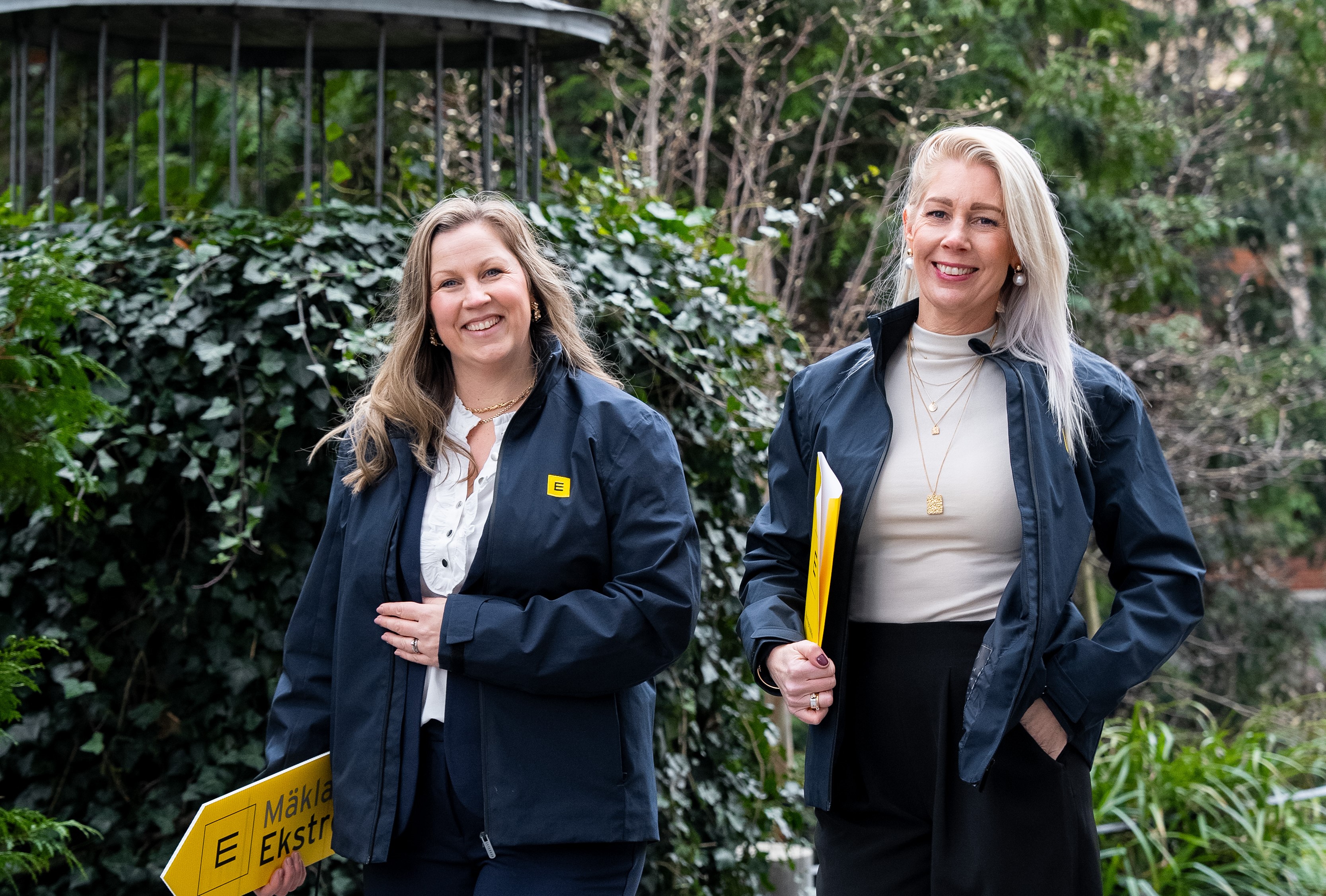 Fastighetsmäklare och kontorschef Frida Stridh, till vänster, och fastighetsmäklare Josefin Nilsson, till höger, går mot kameran och ler. De håller en skylt från Mäklarna Ekström då de ska på visning.